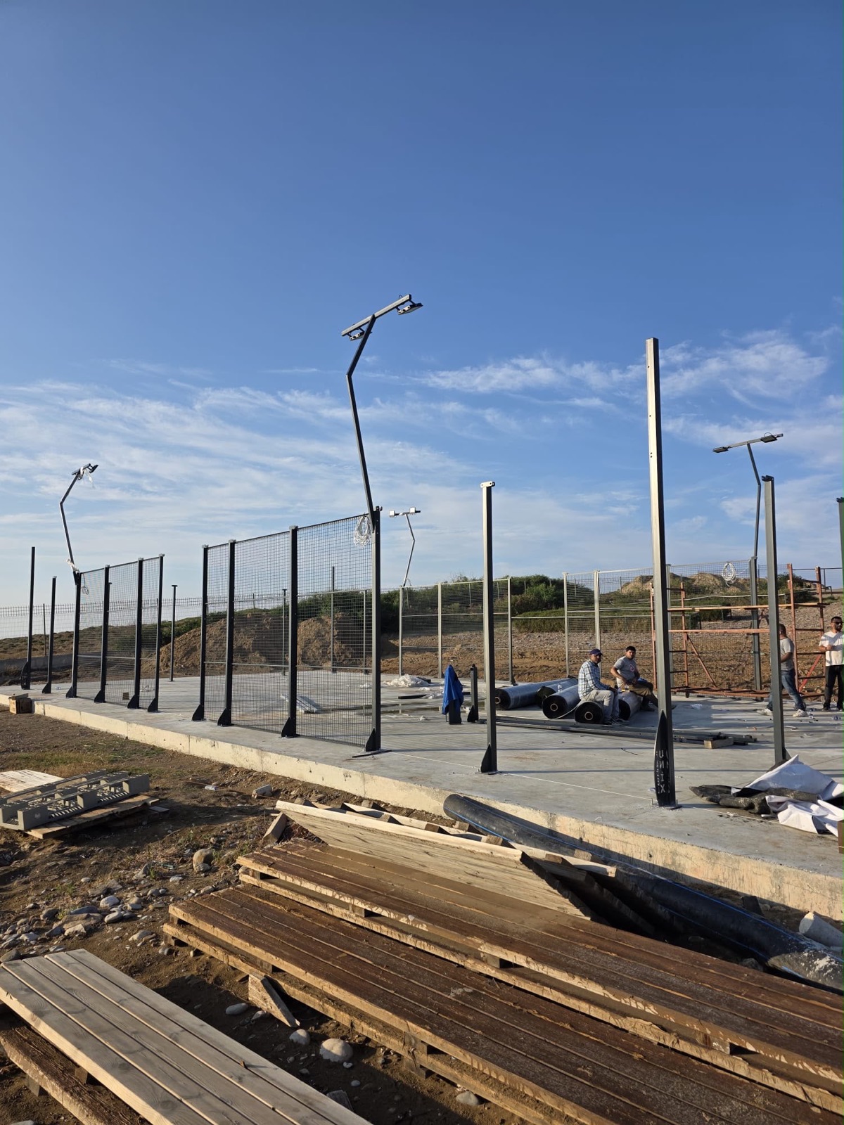 Baustelle mit Betonfundament, montierten Flutlicht-Masten und Montageteam